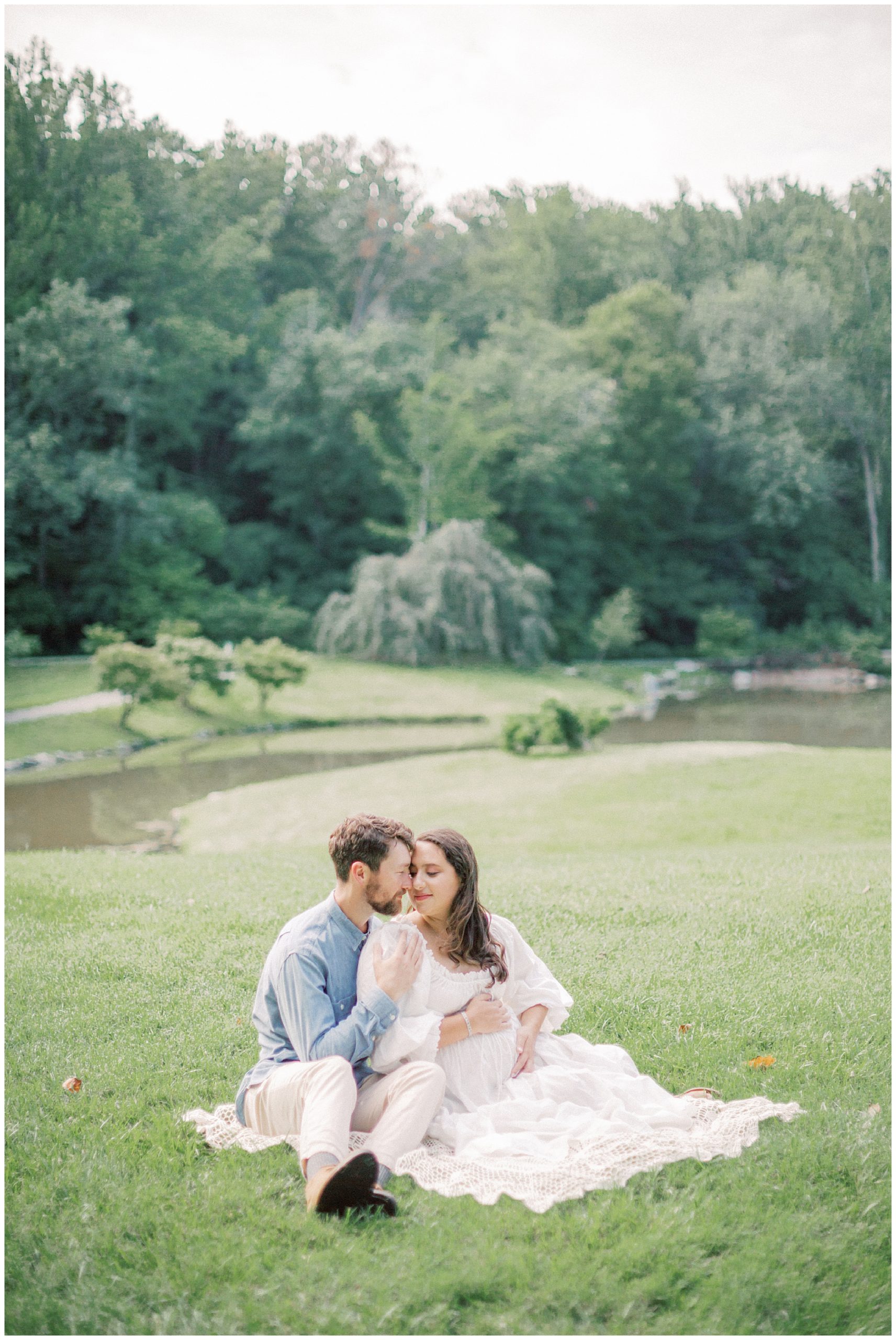 Expecting Father And Mother Sit On Blanket In An Open Lawn During Their Maternity Session.