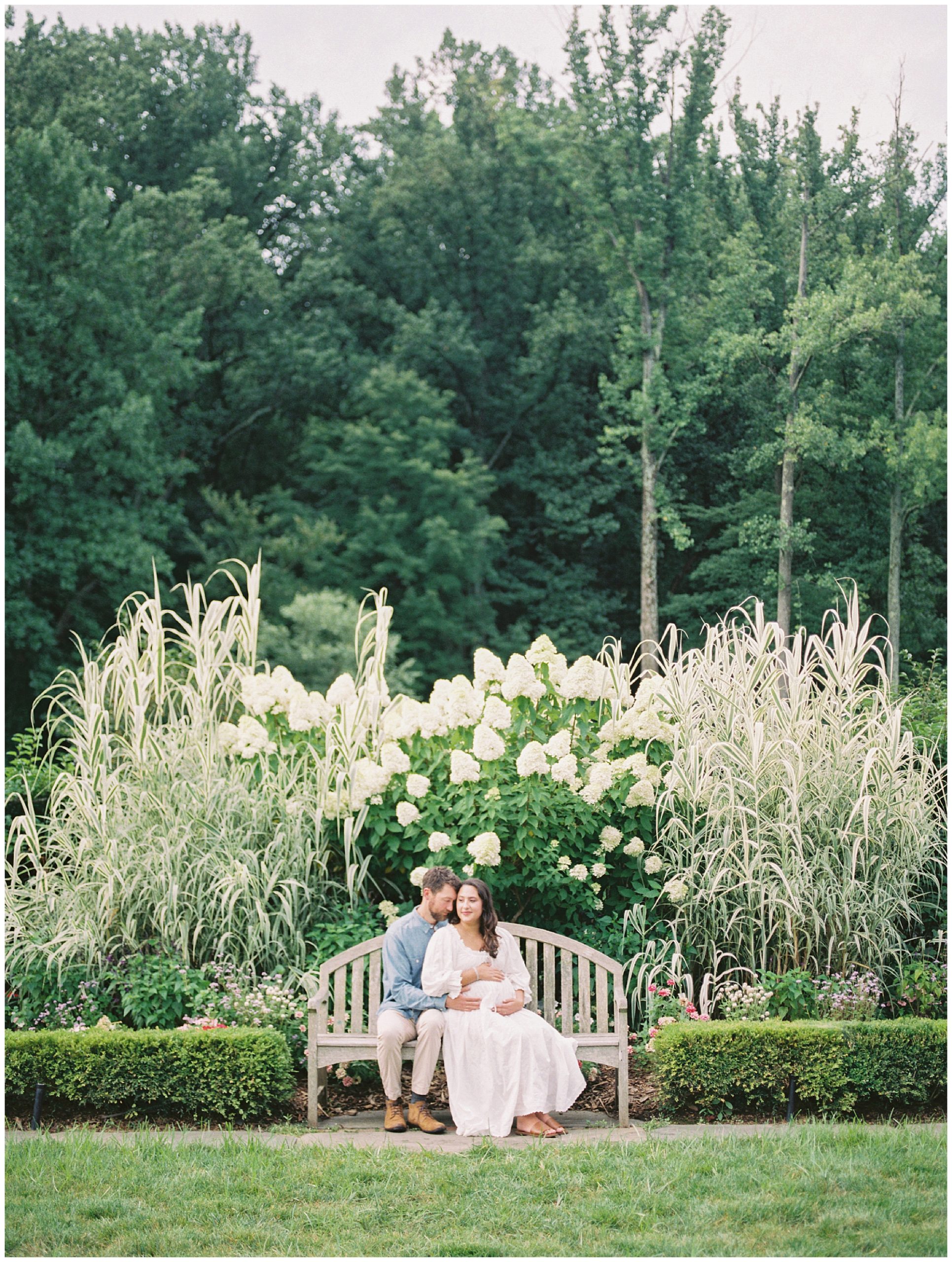 Mother And Father Sit Together On A Bench During Their Maternity Session At Brookside Gardens.