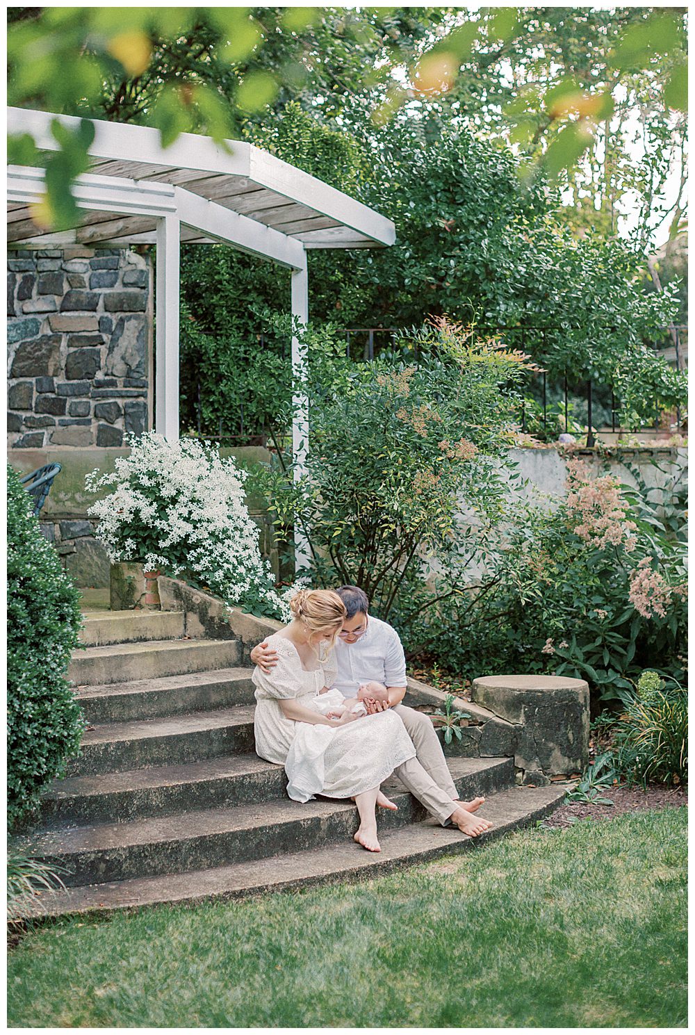 New Parents Sit On Steps In A Garden Holding Their Newborn Baby.