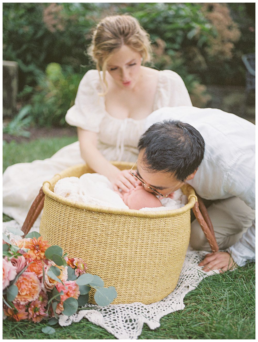 Father Leans Down To Moses Basket And Kisses Newborn Daughter While Mother Helps Calm Her.