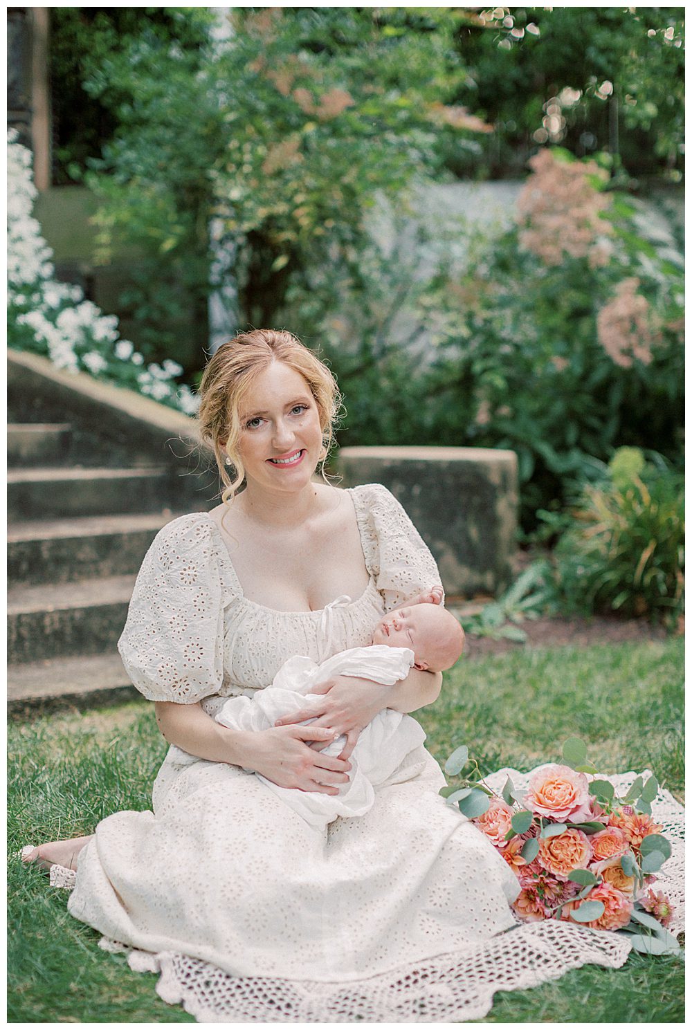 Blonde Mother Sits On Crochet Blanket Holding Newborn Daughter While Smiling At Camera.