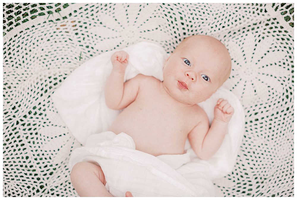 Newborn Baby Looks Up At Camera While Laying On Crochet White Blanket Outside.