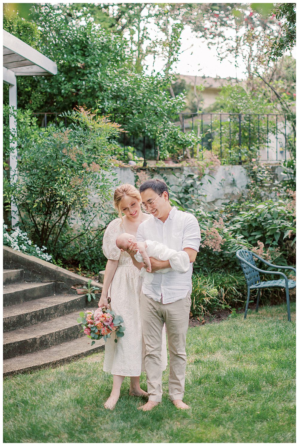 Father Holds Newborn Daughter While Mother Stands With Him Holding Bouquet Of Roses During Outdoor Newborn Session.