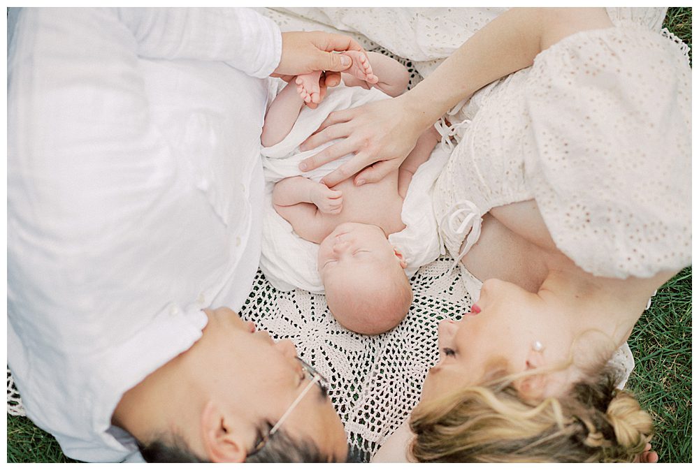 Mother And Father Lay On Crochet Blanket With Newborn Daughter During Their Outdoor Newborn Session In A Garden.