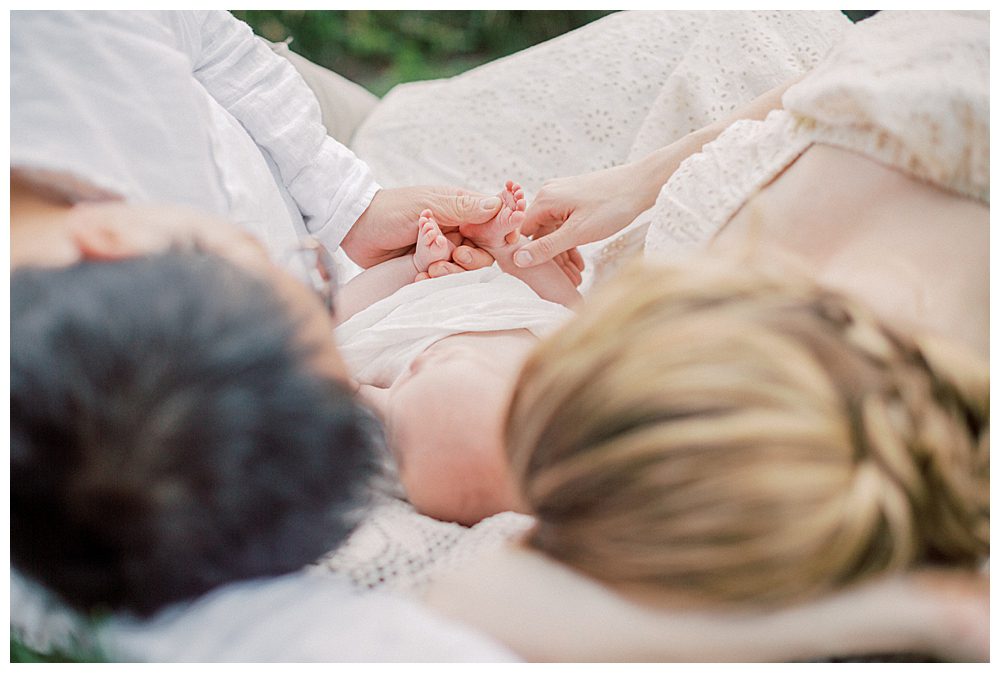 Parents Lay Down On Crotchet White Blanket, Touching The Feet Of Their Newborn Baby Girl.