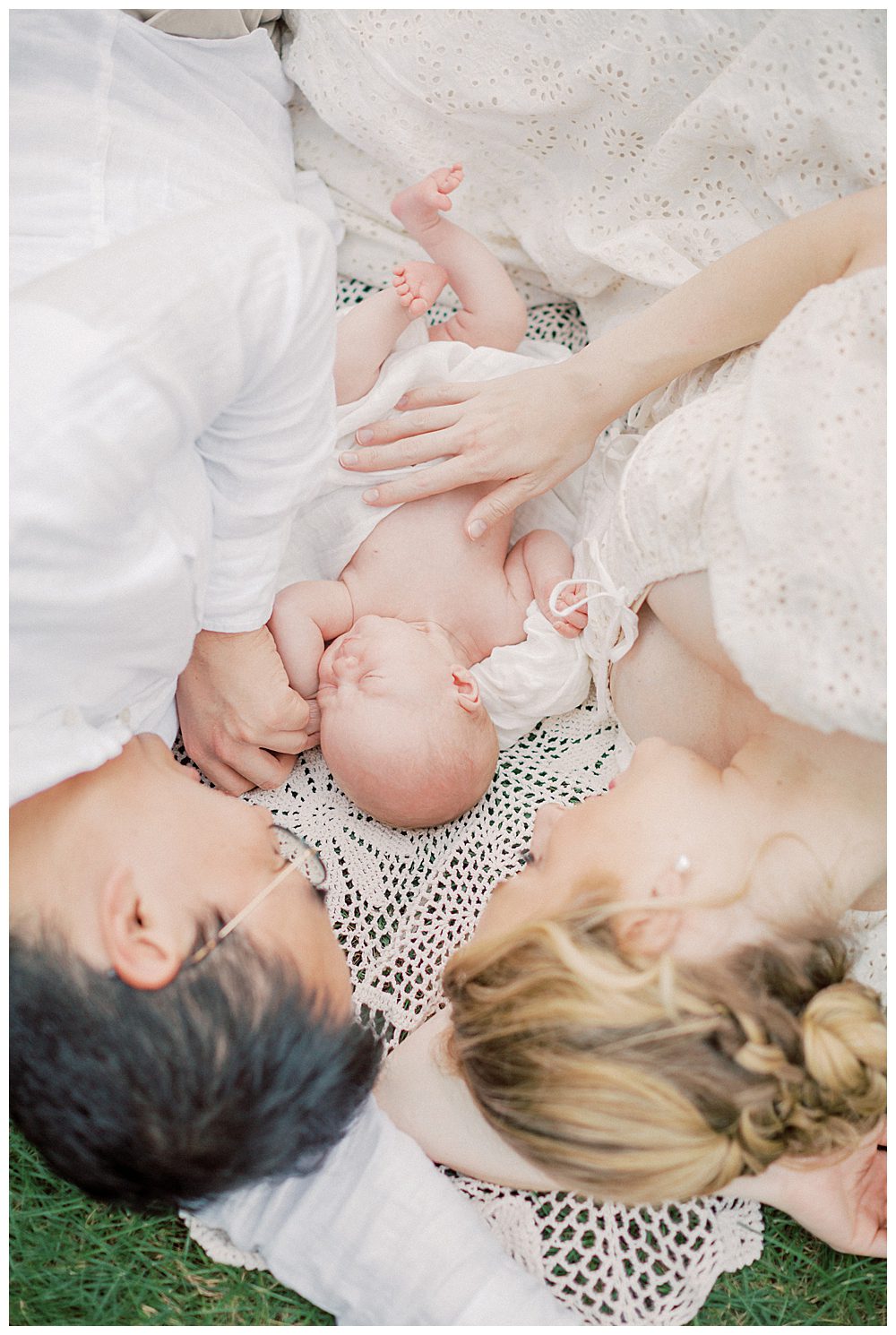 Mother And Father Lay On Blanket Outside With Their Newborn Daughter In-Between Them.