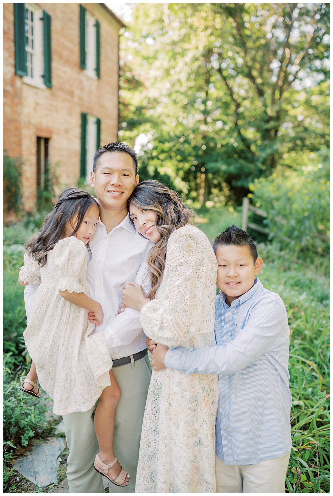 Family Of Four Stand In Front Of Brick House At Colvin Run Mill, Hugging.