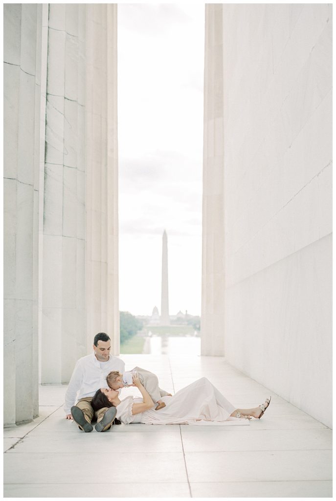 Woman Lays In Husband's Lap While Kissing Baby Boy In The Lincoln Memorial.