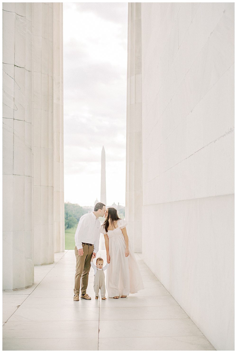 Parents Hold Their Toddler's Hands And Lean In To Kiss While At The Lincoln Memorial.