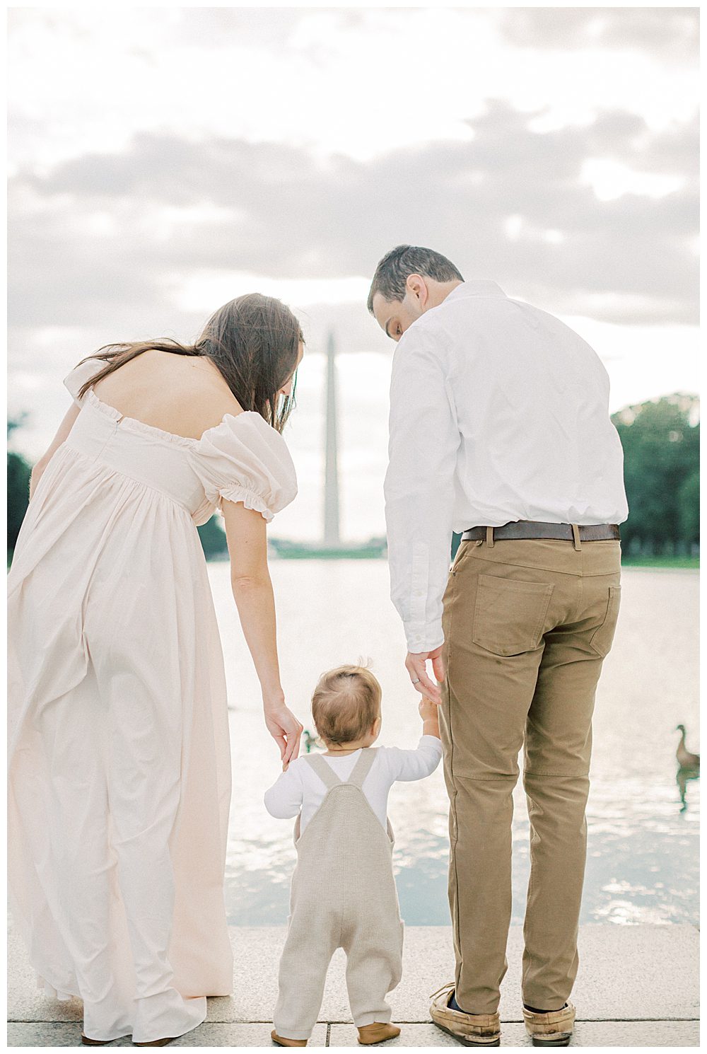 Parents Lean Down And Hold Toddler's Hands While Standing, Looking Out At Dc Reflecting Pool During Dc Monuments Family Photo Session.