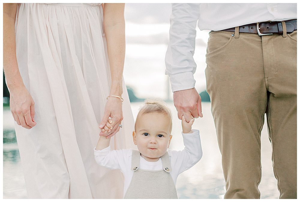 Little Boy Smiles At Camera While Holding Parent Hands.