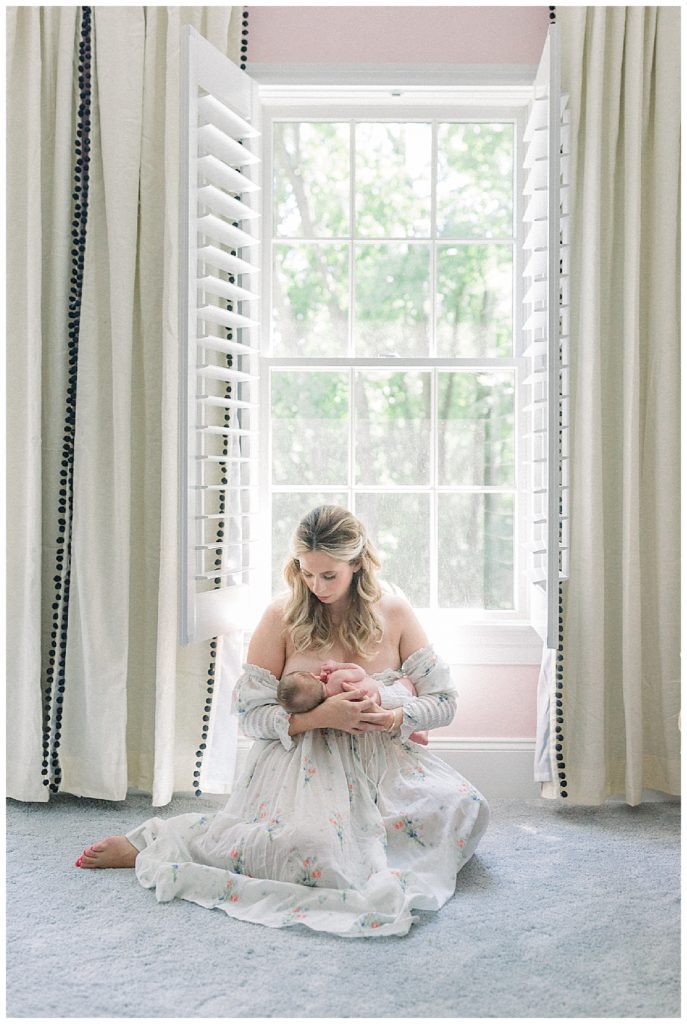 Blonde New Mother Nurses Her Baby Girl While Sitting In Front Of A Large Window, Wearing The Doen Painted Floral Dress.