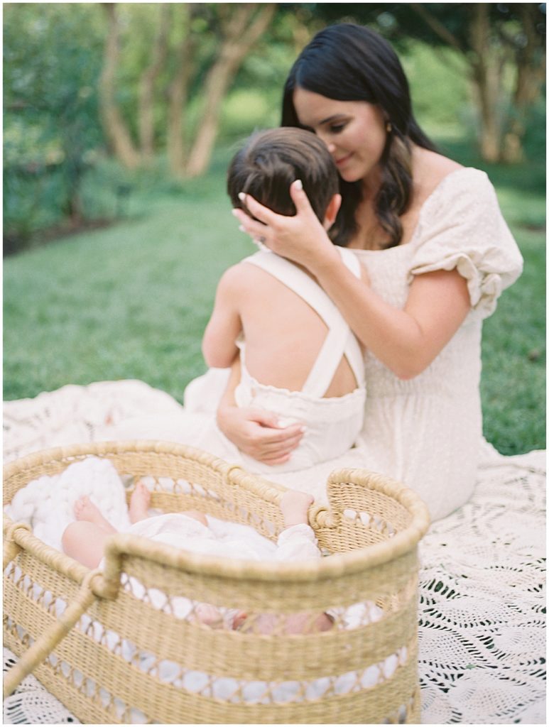 Mother Sits On A Blanket Outside With A Moses Basket, Cradling Her Toddler Son.