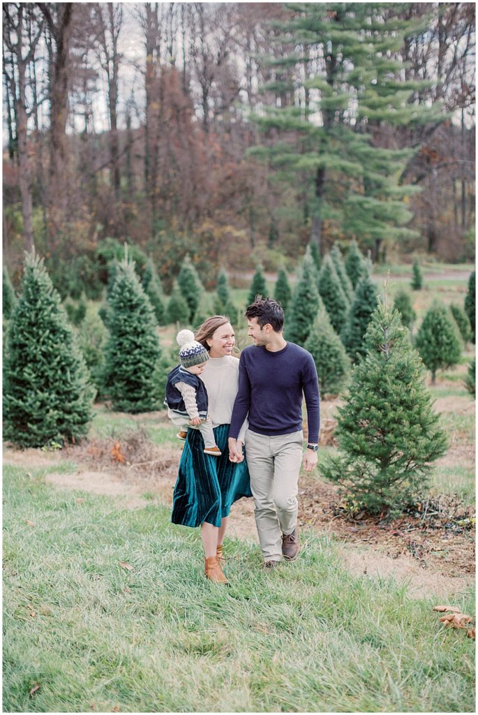 Parents Smile And Walk Holding Their Infant Son During Christmas Mini Sessions At Butler's Orchard.