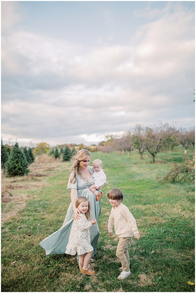 Mom In Green Baltic Born Dress Reaches For Her Children During Christmas Tree Farm Mini Sessions At Butler's Orchard.