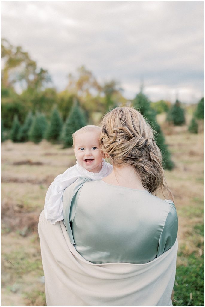 Mom Holds Infant Daughter Over Her Shoulder During Christmas Tree Farm Mini Sessions At Butler's Orchard In The Dmv.