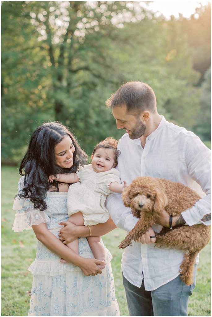 Mother And Father Hold Their Infant Daughter And Goldendoodle Puppy During Montgomery County Family Session At Brookside Gardens..