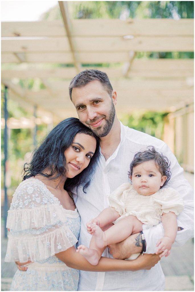 Mother Leans Into Her Husband As They Hold Their Infant Daughter And Look At The Camera During Montgomery County Family Session At Brookside Gardens.