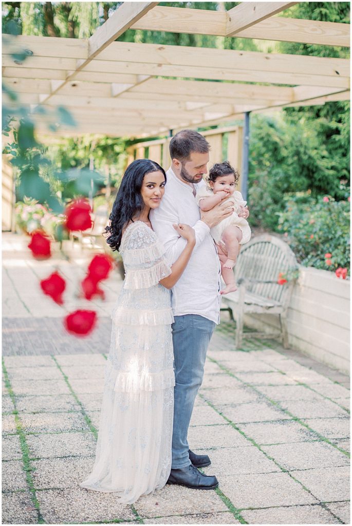 Mother Leans Against Husband As He Holds Their Infant Daughter Under A Floral Pergola During Montgomery County Family Session At Brookside Gardens.