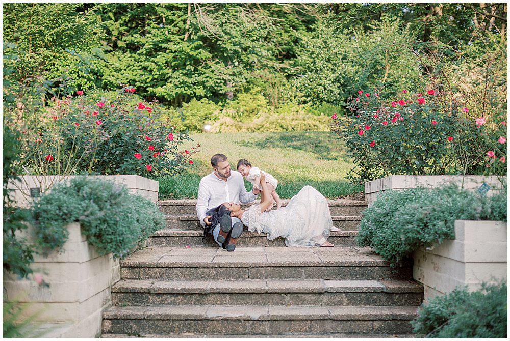 Mother Lays In Husband's Lap While Holding Up Her Infant Daughter As They Sit On Steps At Brookside Gardens In Montgomery County, Maryland.