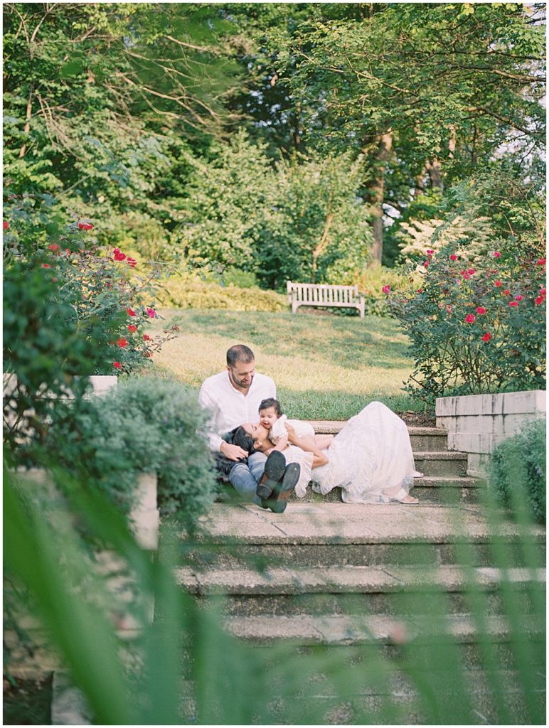 Mother Lays In Husband's Lap And Brings Baby Girl Up To Her Face To Kiss Her During Montgomery County Family Session At Brookside Gardens.