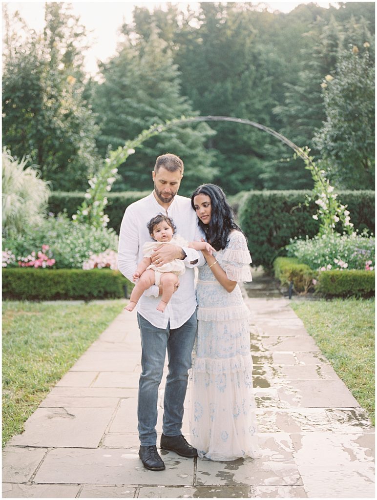 Mother And Father Hold Infant Daughter Near Floral Arch During Montgomery County Family Session At Brookside Gardens.