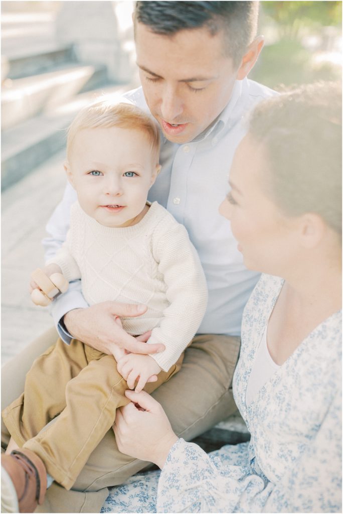 Blonde Little Boy Sits On Parents Lap And Looks At Camera.