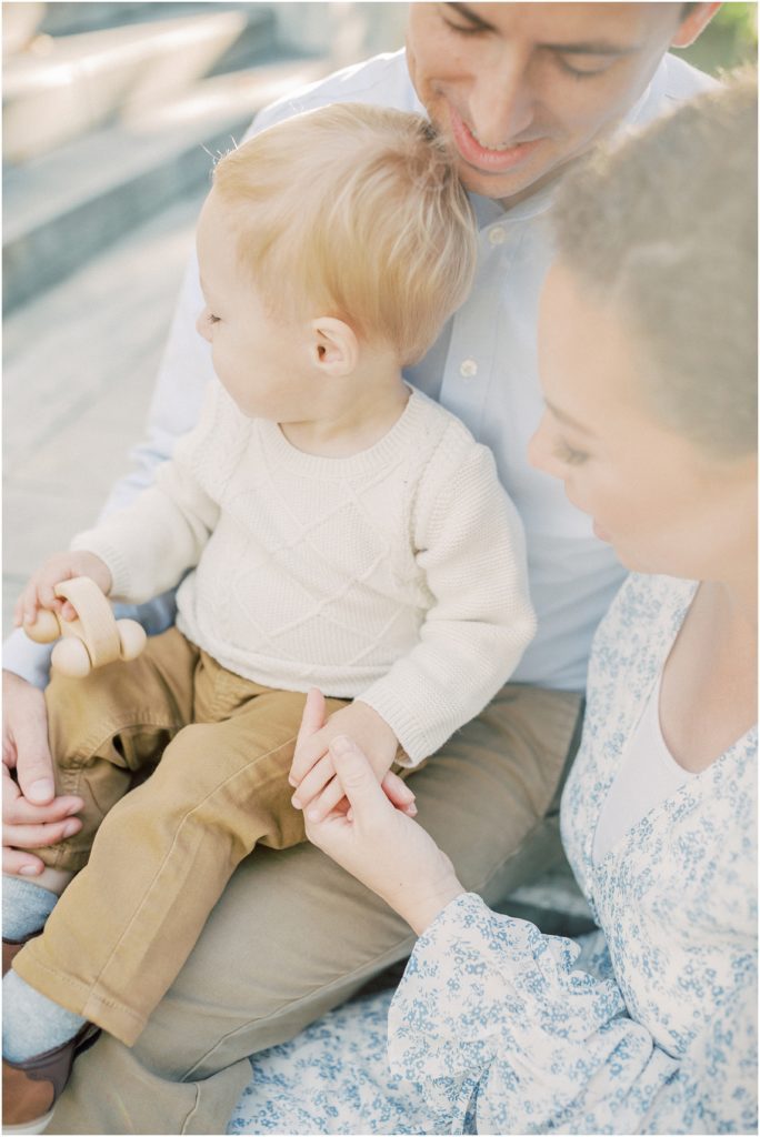 Mother And Father Hold Blonde Son On Their Lap As He Plays With Wooden Car.