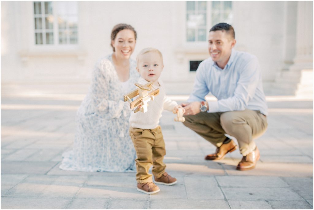 Parents Kneel, Watching Their Toddler Son Play With A Wooden Plane.