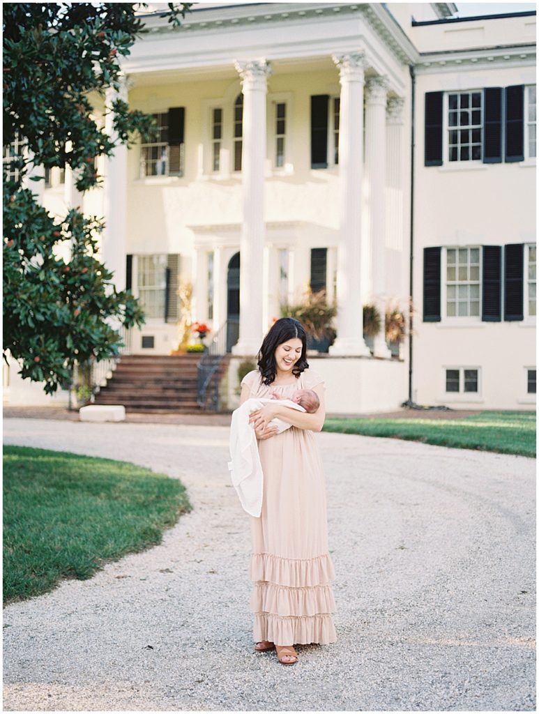 Mother Smiles While Holding Her Infant Son In Front Of Oatlands Historic Home During Newborn Session By Loudoun County Newborn Photographer Marie Elizabeth Photography.