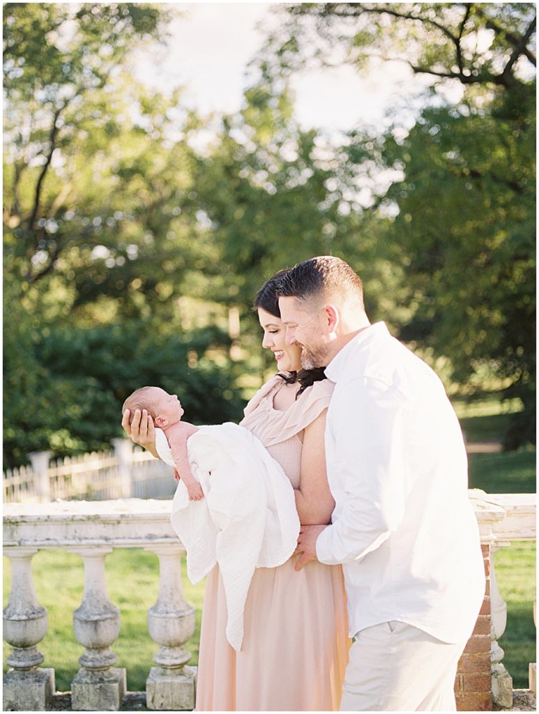 Mother And Father Smile Down As They Hold Their Newborn Son During Outdoor Newborn Session.