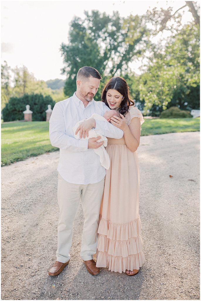 Mother Smiles Down At Newborn Held By Father During Outdoor Newborn Session At Oatlands Historic Home.