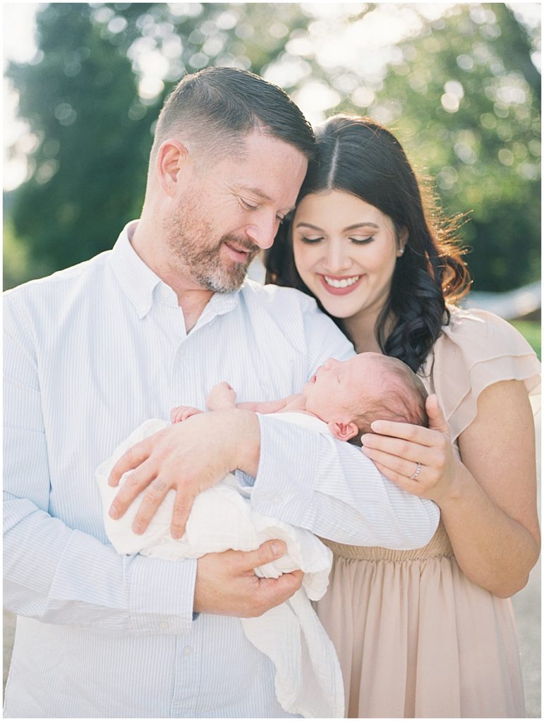 New Parents Hold And Smile Their Baby Boy While Standing Outdoors.