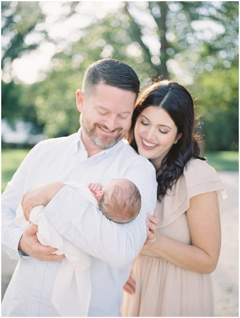 Mother And Father Laugh As They Hold Their Baby Boy Outside.