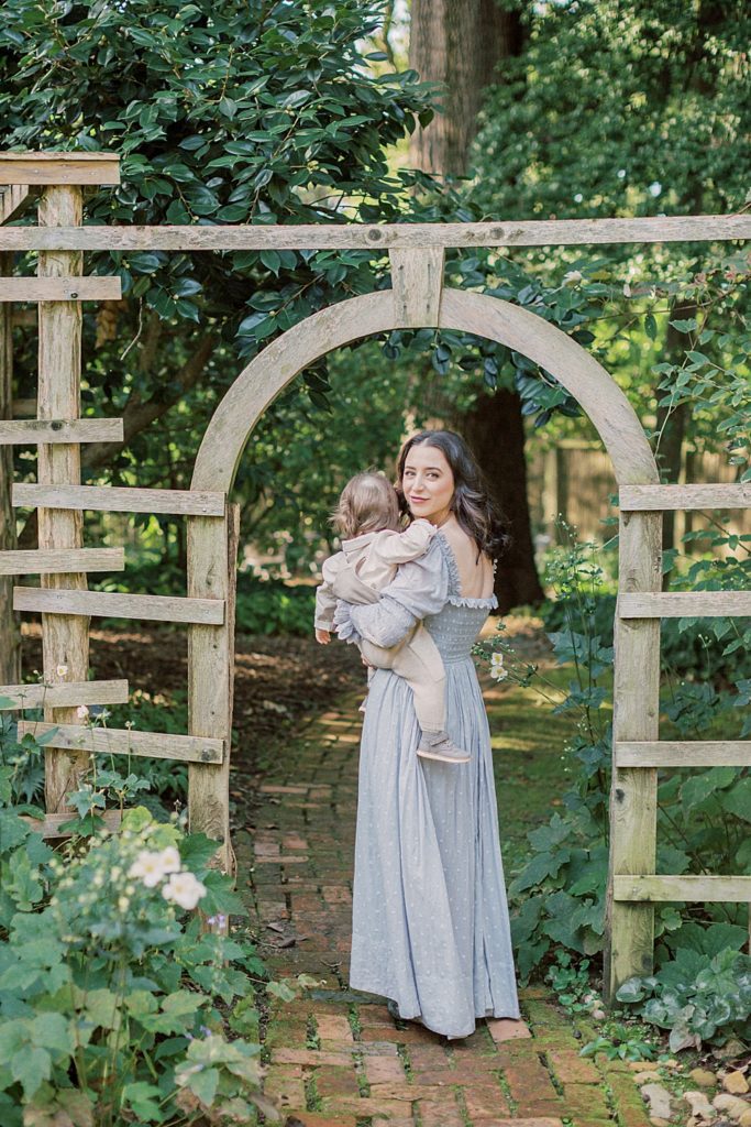 Mother With Dark Hair Stands Under Arch At Tudor Place Holding Her Baby Boy.