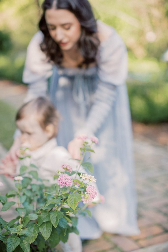 Mother Bends Down To Help Son Walk Around A Garden.