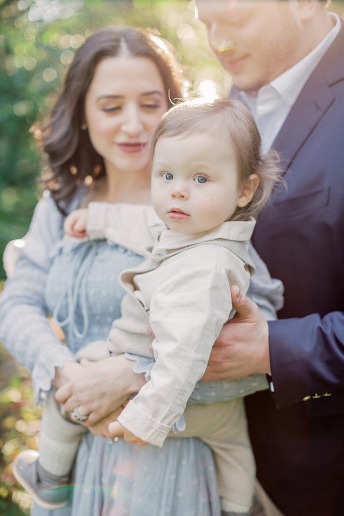 Baby Boy Looks At The Camera With A Soft Smile As He Is Held By His Parents.