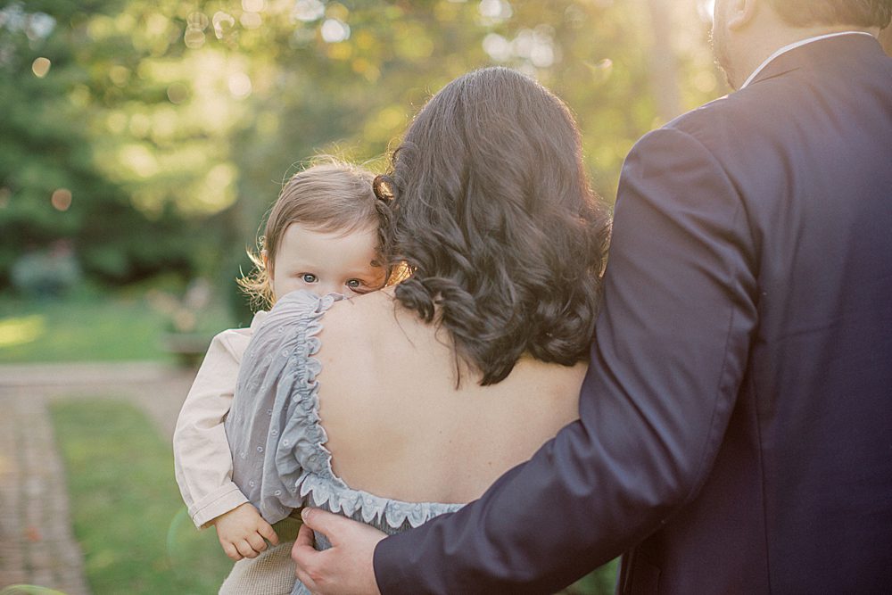 Baby Boy Peeks Over His Mother's Shoulder During Their Family Session.