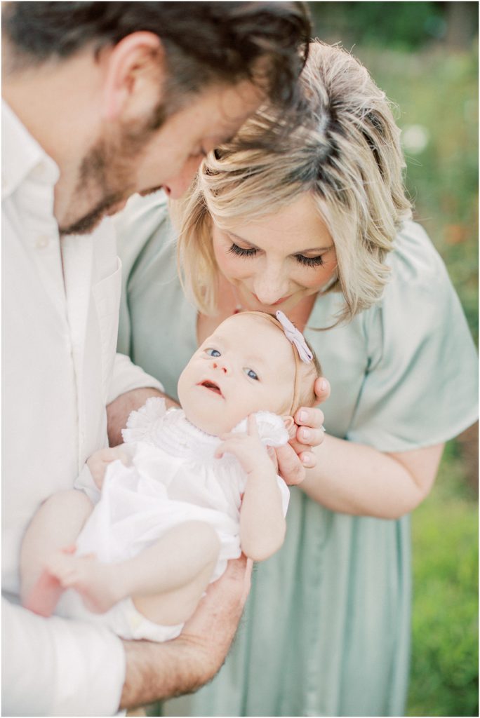 Mother Leans Down And Kisses Baby Girl's Head During Outdoor Newborn Family Photos At Bon Air Rose Garden.