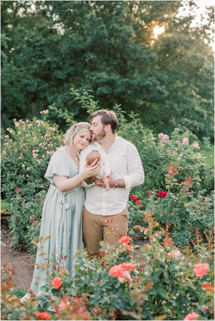 Father Kisses His Wife On The Head As They Hold Baby Girl During Outdoor Newborn Family Photos At Bon Air Rose Garden.