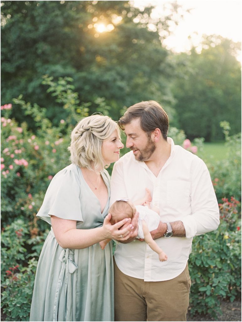 Mother And Father Lean In To Each Other As They Hold Their Baby Girl During Outdoor Newborn Family Photos At Bon Air Rose Garden.