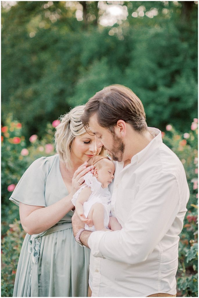 Mother Leans In To Kiss Her Baby Girl On The Head During Outdoor Newborn Family Photos At Bon Air Rose Garden.