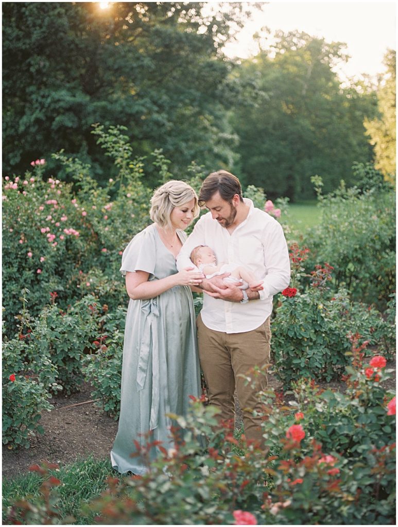 Mother And Father Smile Down At Baby Girl While Standing In Bon Air Rose Garden.