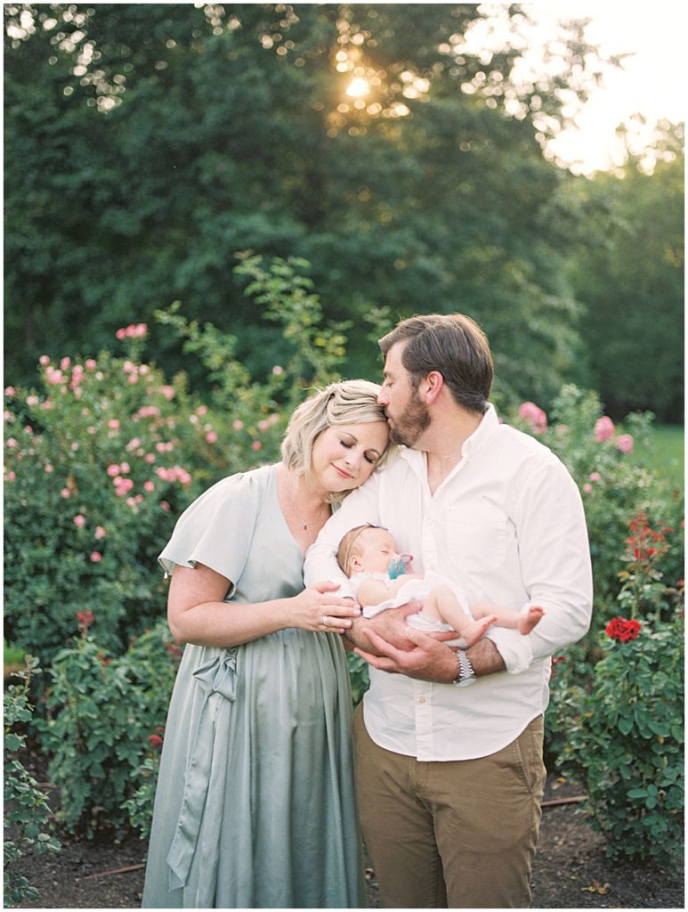 Father Kisses His Wife While Holding Newborn Baby Girl In Rose Garden.