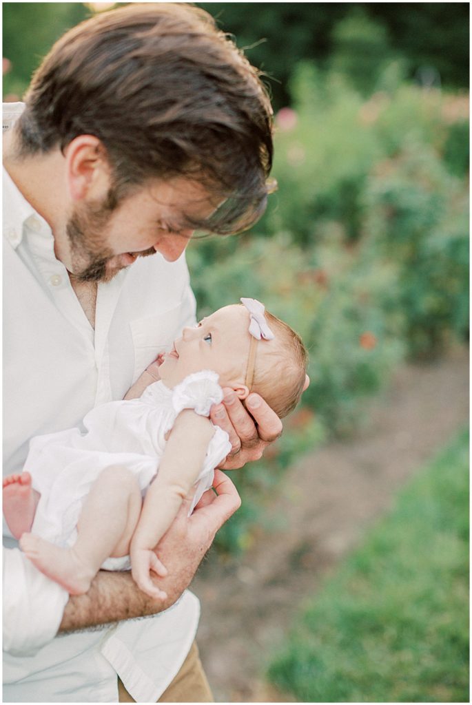 Baby Girl Smiles Up At Her Father During Outdoor Newborn Family Photos At Bon Air Rose Garden.