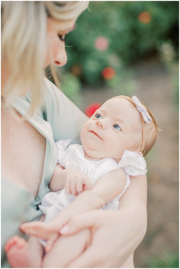 Baby Girl Looks Up At Mom With A Small Smile During Outdoor Newborn Family Photos At Bon Air Rose Garden.