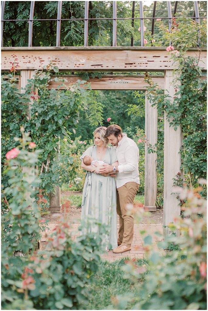 New Parents Stand Together Near Rose Arch During Outdoor Newborn Family Photos At Bon Air Rose Garden.