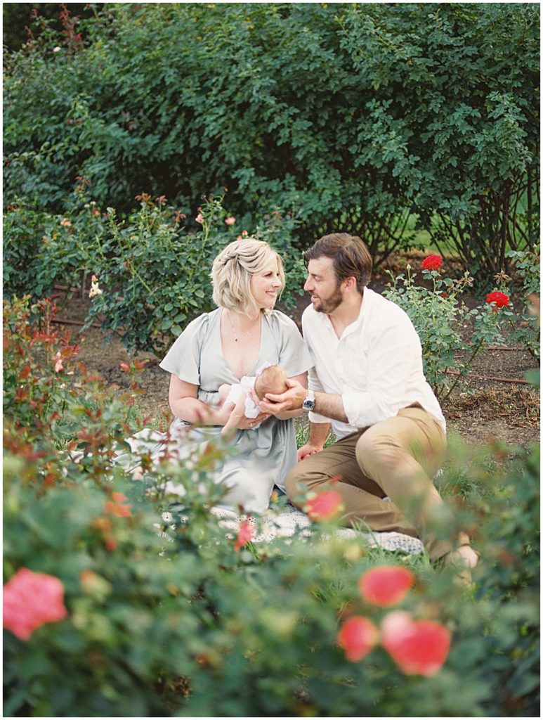 Parents Sit In Garden During Outdoor Newborn Session At Bon Air Rose Garden.