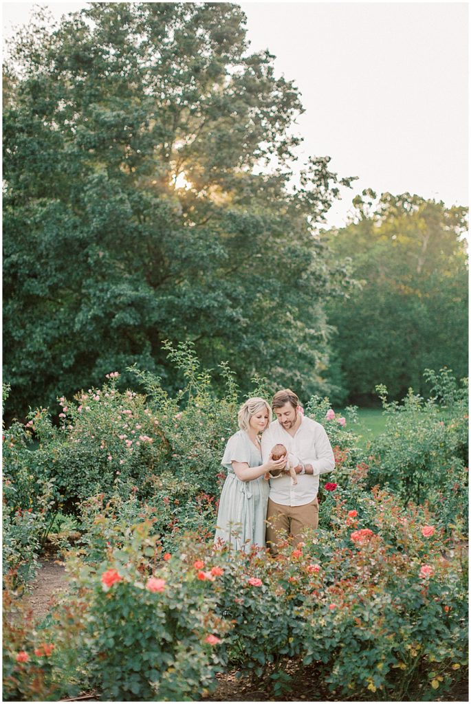 Mother And Father Stand In Bon Air Rose Garden Holding Their Baby Girl During Outdoor Newborn Session.