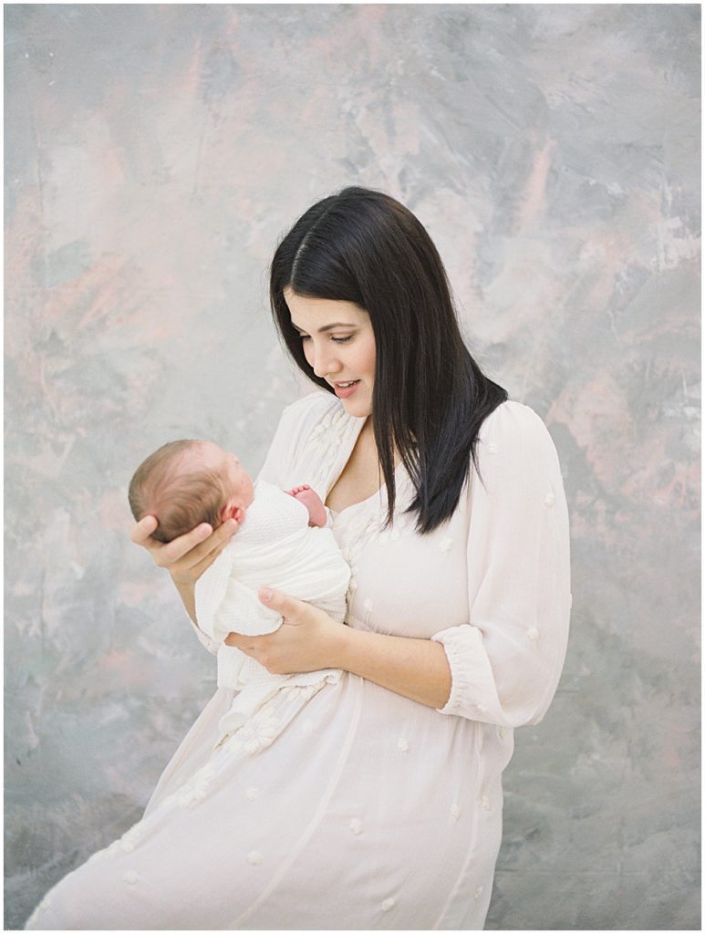 Mother Looks Down At Her Infant Baby Boy Swaddled In White During Studio Newborn Session.